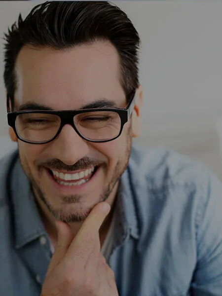A person with glasses and facial hair smiling broadly while resting a hand on their chin.